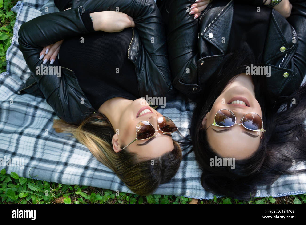 Glückliche Mädchen liegen auf der Decke und Lachen in eine grüne Wiese an einem Frühlingstag in der Natur. Mädchen trägt eine Sonnenbrille und eine schwarze Jacke Stockfoto