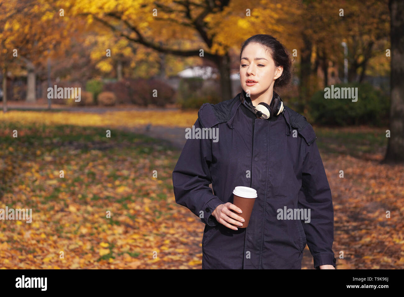 Ehrliche junge Hipster Frau genießen Spaziergang durch Park im Herbst Saison holding Kaffee Becher in der Hand zu gehen und die Kopfhörer um den Hals Stockfoto