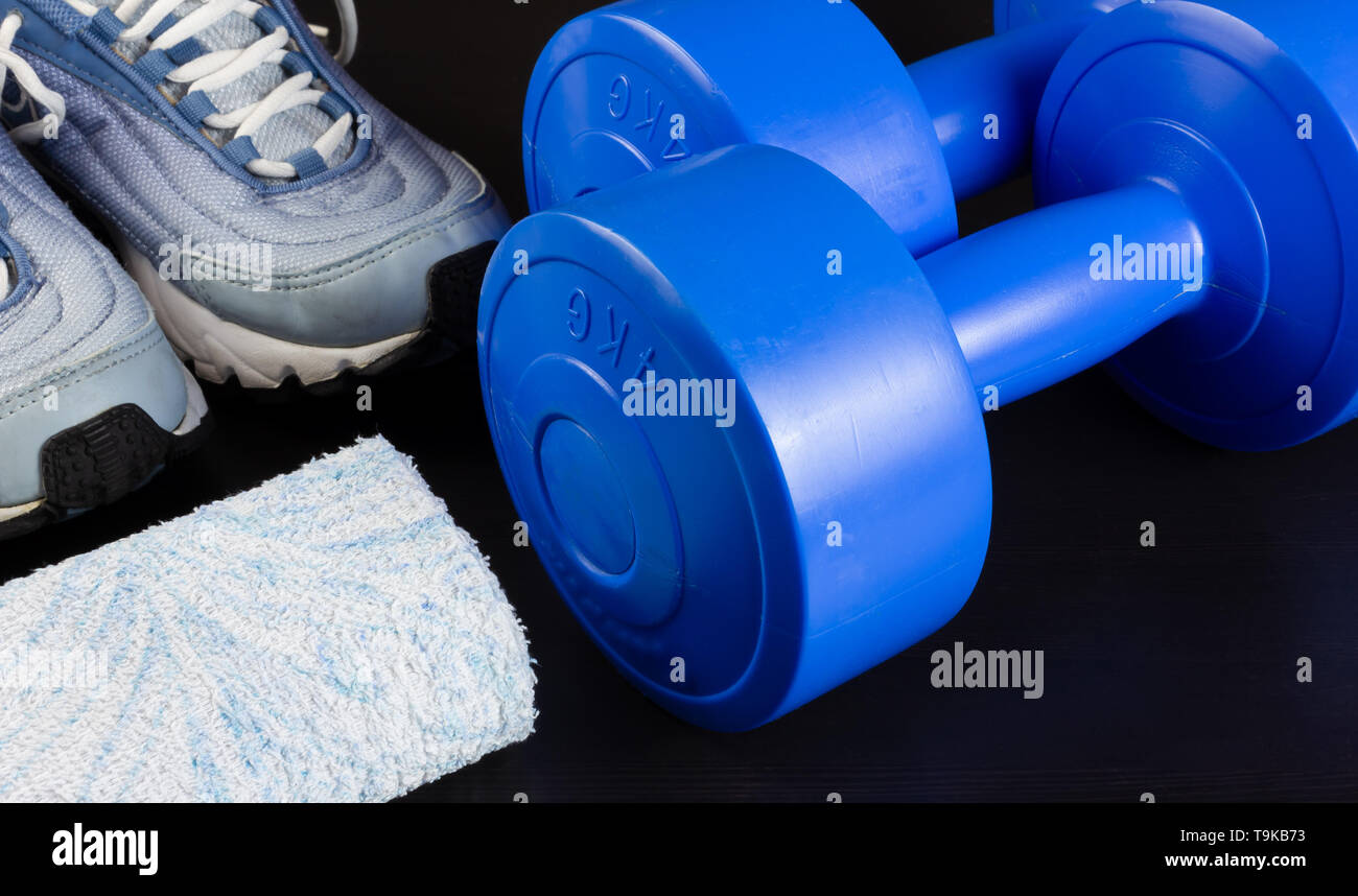 Ein Paar Turnschuhe, zwei blaue Kurzhanteln und ein Handtuch auf einem dunklen Hintergrund Stockfoto