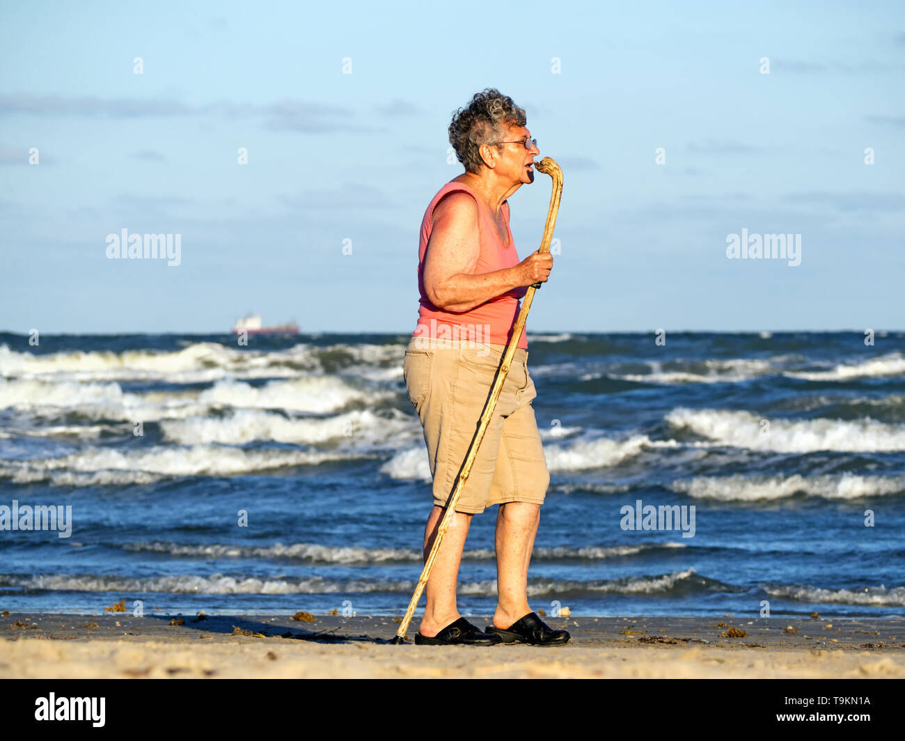 Eine ältere kaukasische Frau mit einem Stock Spaziergänge neben dem Golf von Mexiko am Strand während der Goldenen Stunde in Port Aransas, Texas USA. Stockfoto