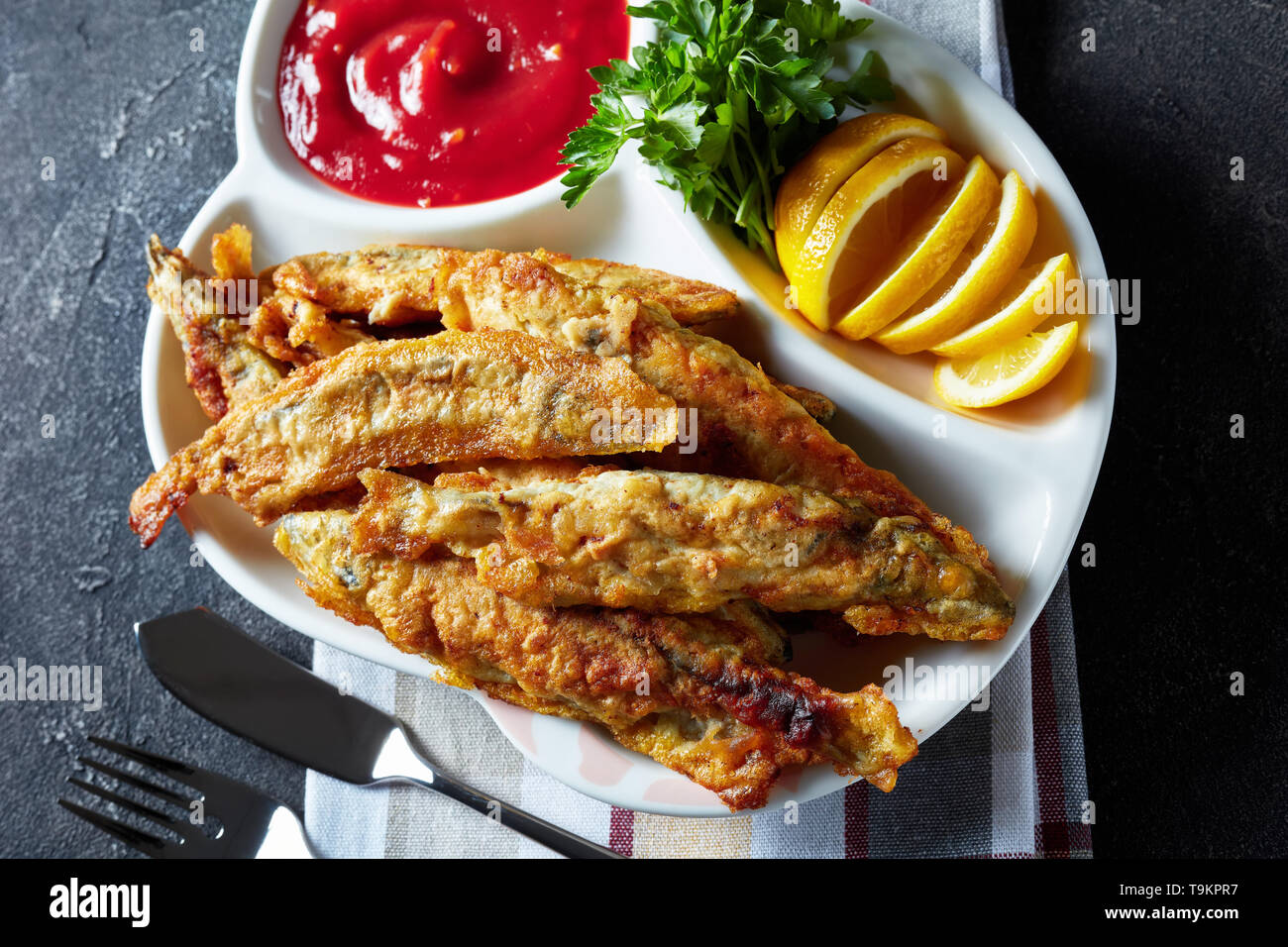 Gebratene kleine capelins mit Tomatensauce, in Scheiben geschnittenen Zitrone auf einer Platte auf einen konkreten Tisch mit Messer und Gabel, Ansicht von oben, flatlay, close-up Stockfoto