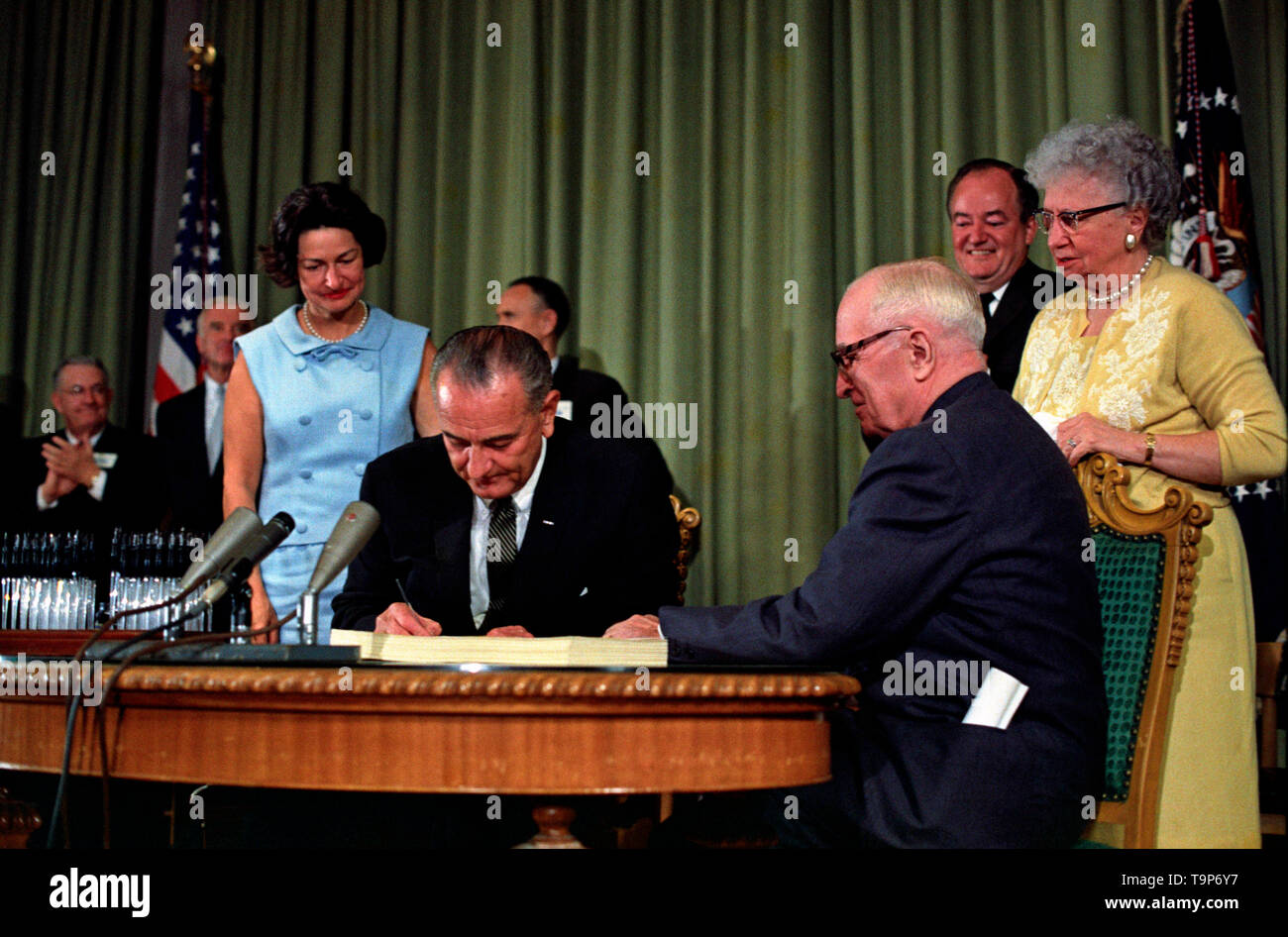 Präsident Lyndon B. Johnson die Unterzeichnung der Medicare Rechnung am Harry S. Truman Library in der Unabhängigkeit, Missouri. Der ehemalige Präsident Harry S. Truman ist am Tisch mit Präsident Johnson sitzt. Die folgenden sind im Hintergrund (von links nach rechts): Senator Edward V. Lange, ein unbekannter Mann, Lady Bird Johnson, Senator Mike Mansfield, Vizepräsident Hubert Humphrey, und Bess Truman. Juli 30, 1965 Stockfoto