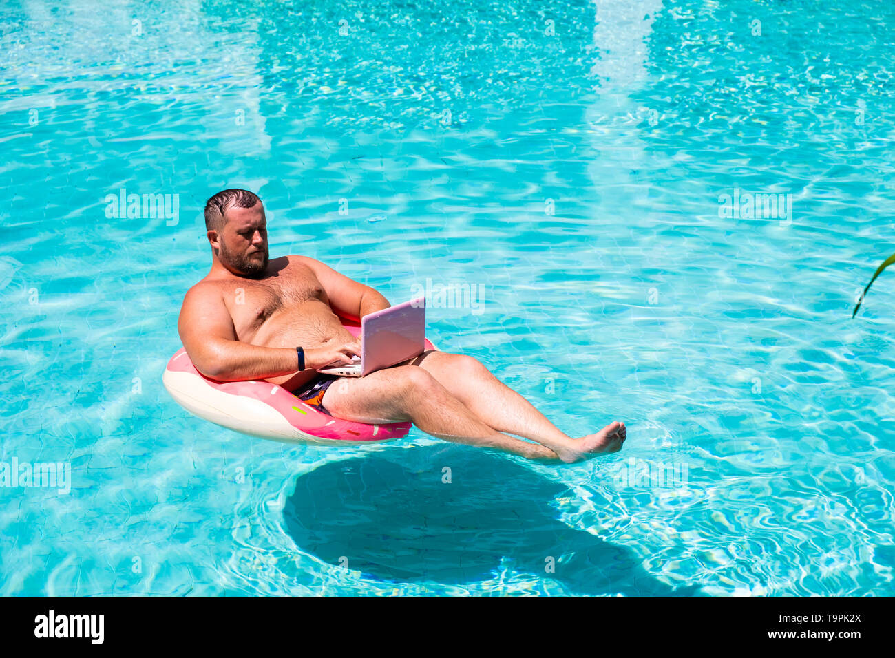 Mann mit einem aufblasbaren Ring in das Wasser im Pool arbeitet auf dem Computer. Funny Fat Man in einen Kreis mit pinkfarbener Laptop. Freiberufliche Konzept Stockfoto