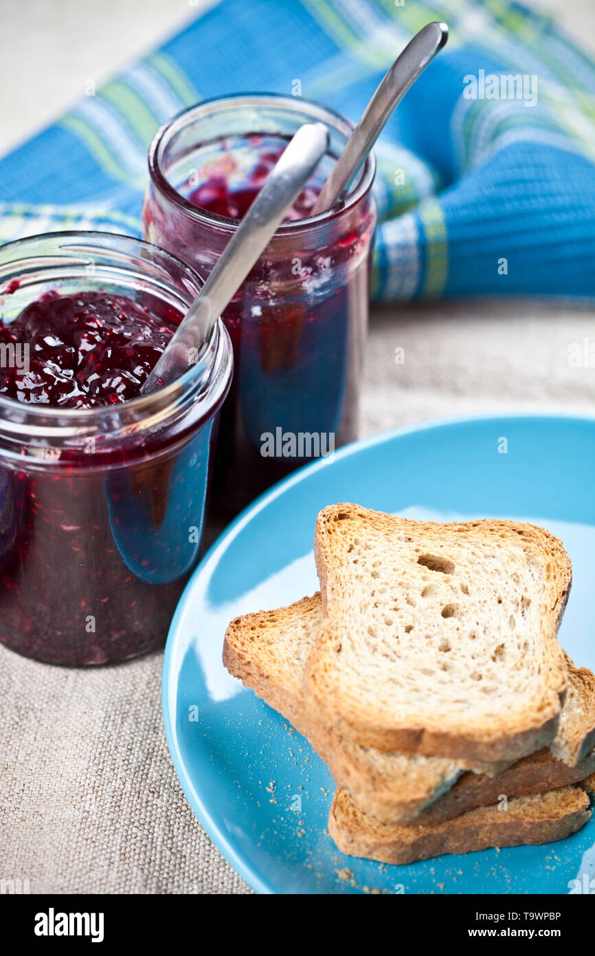 Geröstetes Getreide Brotscheiben auf blau Keramik Platte und hausgemachte Kirsche und Beeren Marmelade in Gläser und Löffel closeup und Bettwäsche Serviette auf rustikalen Woode Stockfoto