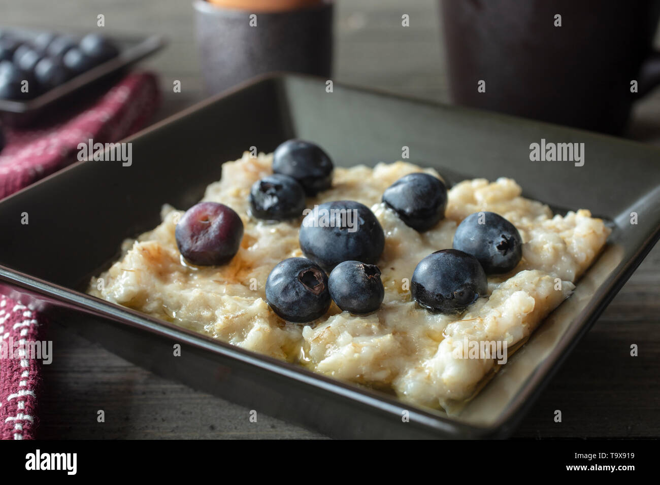 Frühstück Schüssel Haferflocken, Honig, Butter und Blaubeeren mit Kaffee und weich gekochte Eier auf einem rustikalen Tisch mit einem Löffel und Geschirrtuch Stockfoto