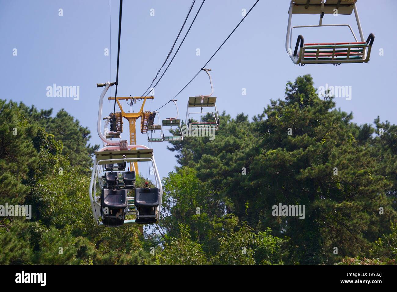 Seilbahnen durch Baumkronen - Skilift Sitze gegen grüne Blätter mit blauen Himmel hinter Stockfoto