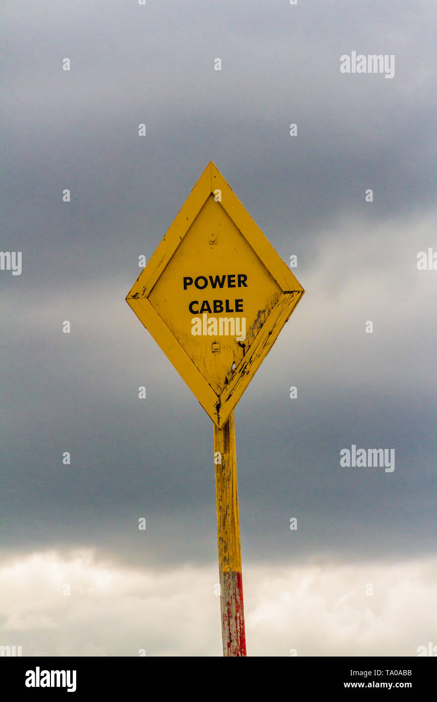 Gelbe Stromkabel Warnzeichen vor einem stürmischen Himmel. Stromversorgung Kabel verläuft zwischen der heiligen Insel von Lindisfarne und dem Festland, Großbritannien. 2018. Stockfoto