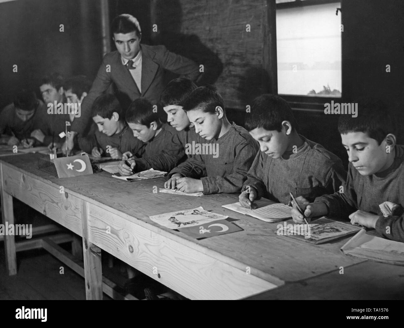 Foto der Türkischen jungen Schreiben und Malen im Klassenzimmer einer Schule, während ein Lehrer ist der Überprüfung ihrer Arbeit. Auf der langen Schreibtische gibt es kleine Bilder von der Flagge der Republik Türkei. Die neue türkische Schrift mit lateinischen Zeichen hatte für die Kinder in allen Schulen unterrichtet seit der Jahreswende 1928/1929. Stockfoto