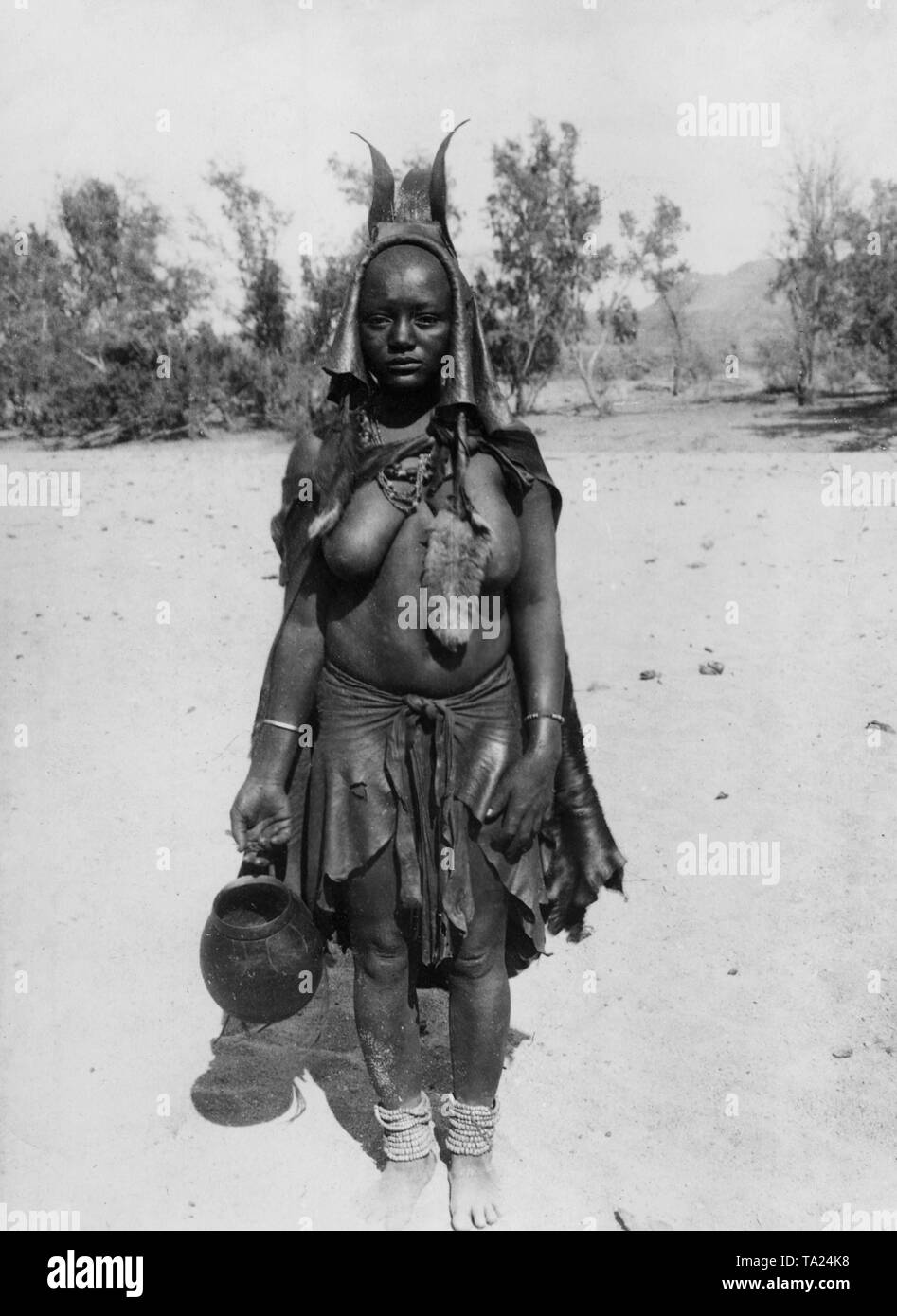 Herero Frau in Tracht, mit Kopftuch und Gefäß in der Hand auf einem Sand Oberfläche in Deutsch-südwestafrika (undatierte Aufnahme). Stockfoto