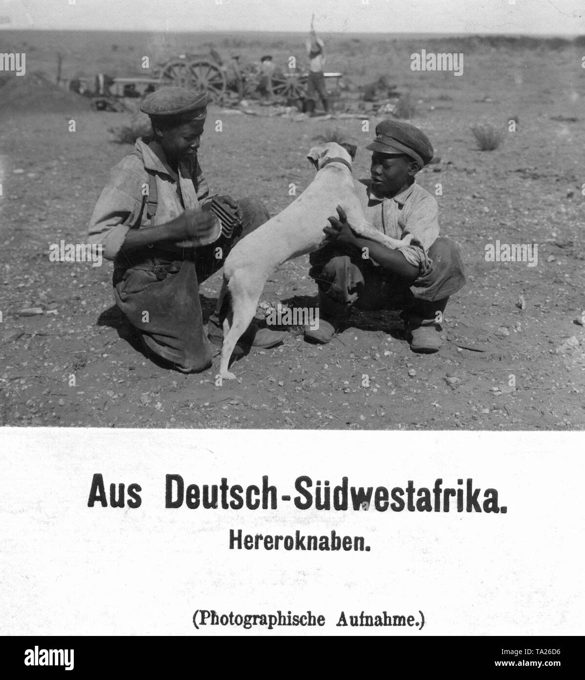 Zwei Jungen, die von den Herero im Europäischen Kleidung spielen sie mit einem Hund in das Feld "Öffnen" ein. Im Hintergrund die Männer arbeiten. In Deutsch-südwestafrika (Undatiertes Foto). Stockfoto