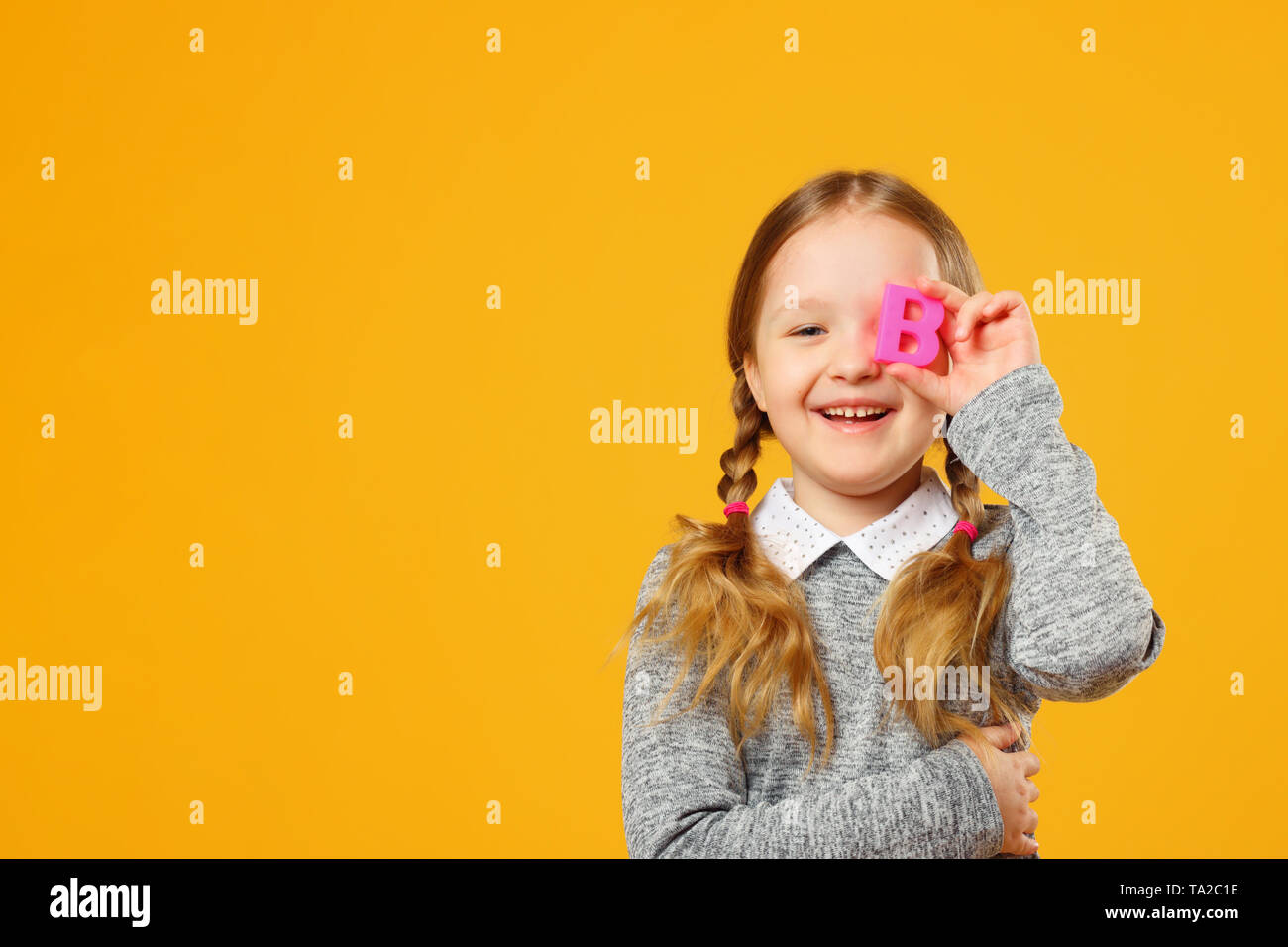Portrait von Eine fröhliche kleine Kind Mädchen auf einem gelben Hintergrund. Schülerin hält der Buchstabe b. Der Begriff der Bildung. Zurück zu Schule. Stockfoto