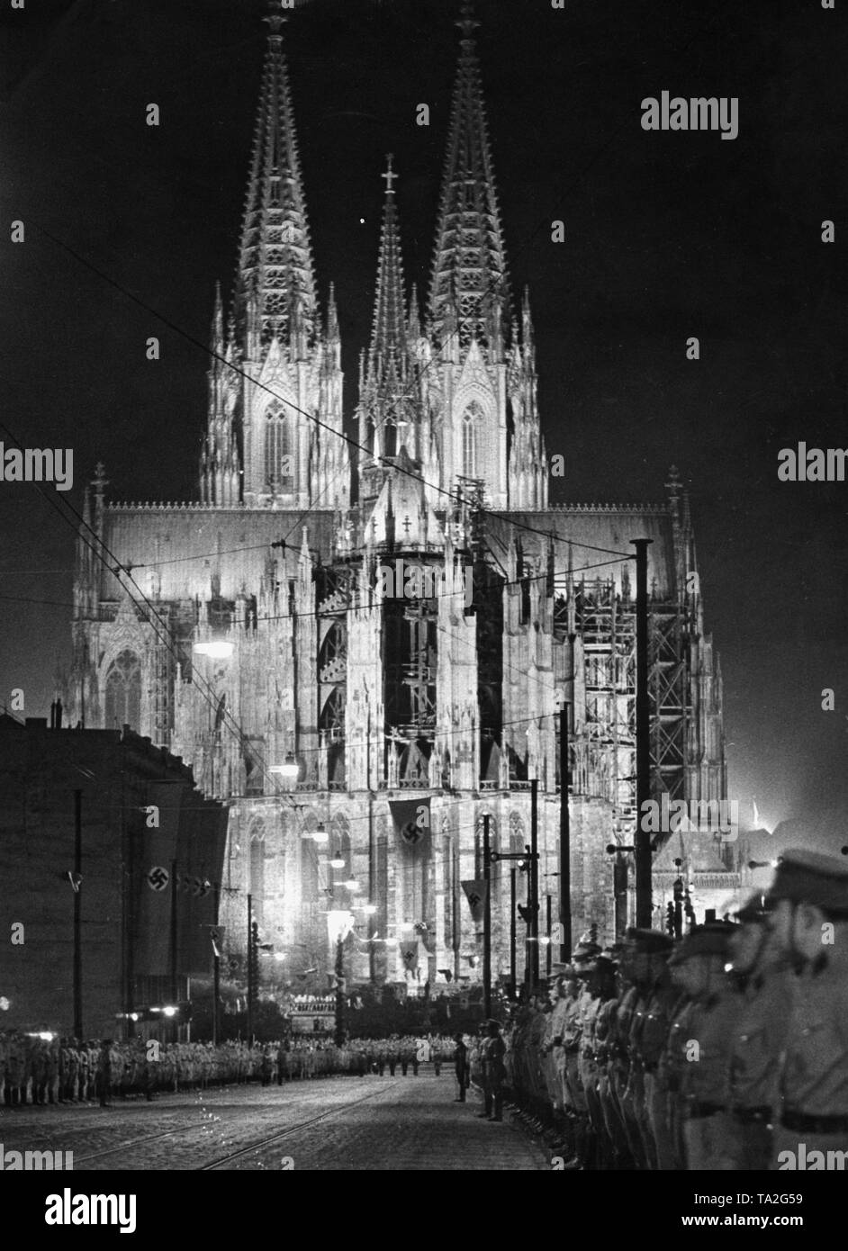 Eine SA März vor dem Kölner Dom am 29.3.1936 nach der Besetzung und die Remilitarisierung des Rheinlandes von Deutschland. Die Kuppel ist mit Strahlern beleuchtet. Uniformierte SA-Mitglieder links und rechts der Straße. Hakenkreuzfahnen hängen von den Laternen und Gebäuden. Stockfoto