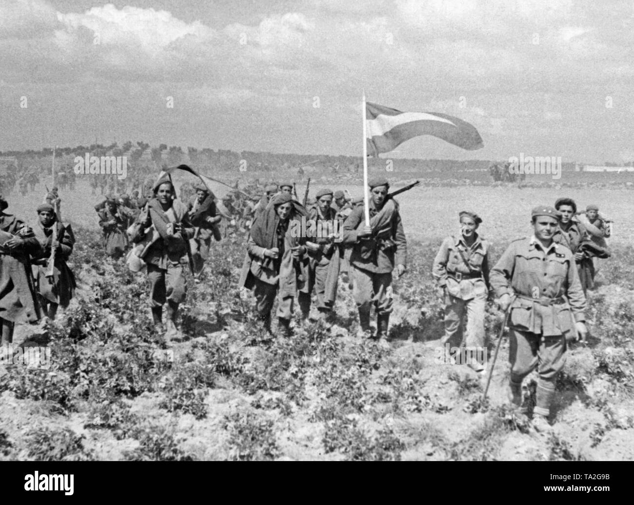 Foto von nicht-regulären nationalen spanischen Truppen marschieren über ein Feld in der Spanischen Meseta in den letzten Tagen des Krieges am 28. März 1939. Die Soldaten tragen ihre gesamte Feld Pack und spanischen nationalen Flaggen (rot-gelb-rot). Im Vordergrund, ein Lieutenant (mit einem Stock und einer Zigarette) Führt die Truppen. Im Hintergrund und Olivenbäumen. Stockfoto