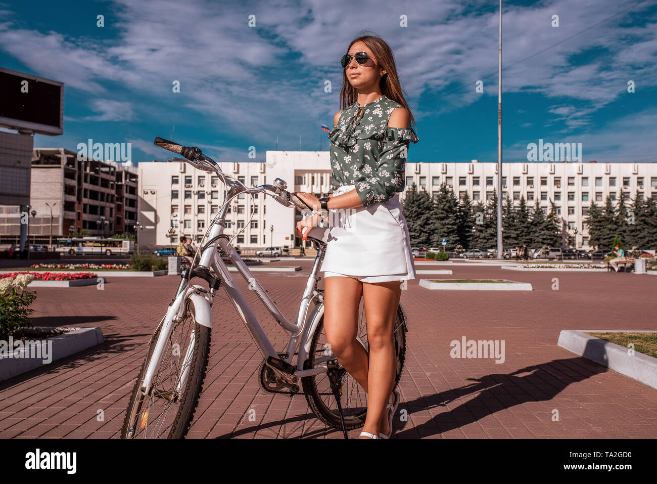 Frau mit einem Fahrrad, Sommer in der Stadt. Hintergrund Gebäude Himmel.  Grüne Jacke und weißen Rock Shorts. Lange Haare Sonnenbrille. Spaziergang  auf dem Fahrrad nach Stockfotografie - Alamy