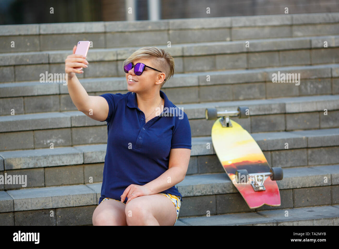 Stilvolle Mädchen von Plus size, modischer Haarschnitt im Mirror Gläsern, sitzen auf der Treppe, Fotos von sich selbst mit einem Smartphone, einem longboard neben Stockfoto