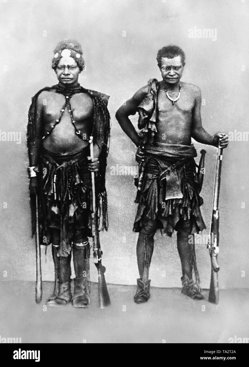 Herero chief Kambazembi wa Kangombe, ein Stammesführer der Herero, der Mann rechts neben ihm unbekannt ist. Beide tragen traditionelle Kleidung, Schmuck und Waffen (undatiert). Stockfoto