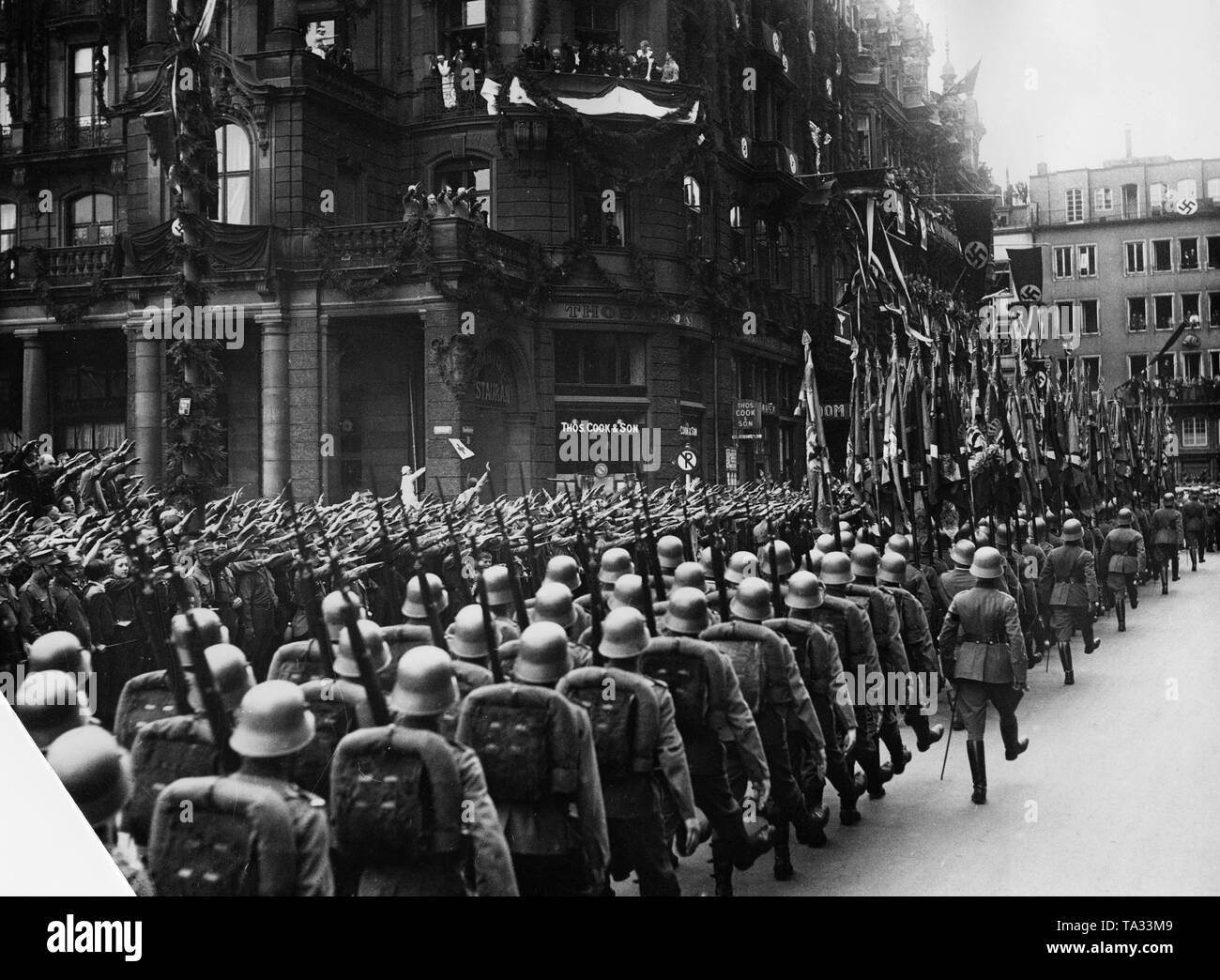 Szene einer militärischen Parade vor dem Dom-Hotel in Köln vom 29.3.1936 nach der Besetzung und die Remilitarisierung des Rheinlandes von Deutschland. In der Mitte, Wehrmachtssoldaten. Weiter vorne im Zug die Fahnen der Rheinischen Regimenter. Links, Thomas Cook Reisebüro. Auf den unteren Balkon des Dom-hotels: Von links: Werner von Fritsch, Robert Ley, Adolf Hitler und Werner von Blomberg. Stockfoto