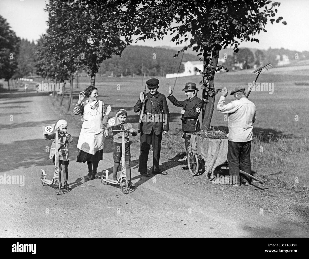 Frau mit Kindern Begegnungen NS-Patrouillen auf der Strasse. Die Kinder haben hakenkreuzfahnen auf ihren Rollern befestigt. Die bewaffneten Männer tragen Uniformen und Hakenkreuz Armbinden. Undatiertes Foto, um 1938. Stockfoto