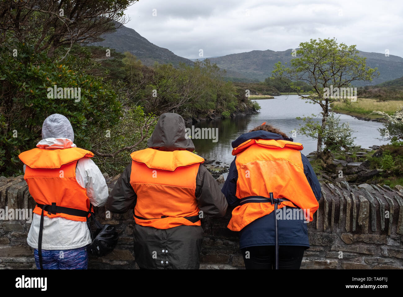 Drei Personen auf einer Brücke, Schwimmwesten, während sie für ein Boot in der Sitzung des Wasser, Ring of Kerry, Irland, an einem bewölkten warten warten Stockfoto