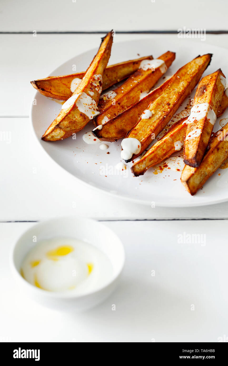 Hausgemachte karamellisierten geröstet geschnitten Süßkartoffeln oder Süßkartoffel mit griechischen Stil Joghurt und zerdrückten roten Pfeffer Flocken close-up auf weißem Holz- Hintergrund. Er Stockfoto