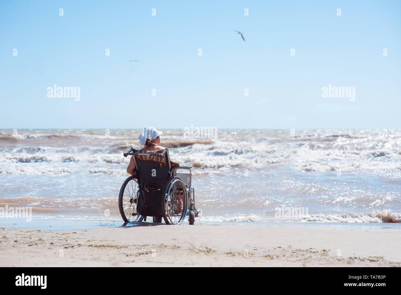 Rückansicht einer behinderten Frau, die an einem sonnigen Tag auf einem Rollstuhl am Strand sitzt. Sie schaut auf den Ozean Stockfoto Rückansicht einer behinderten Frau, die an einem sonnigen Tag auf einem Rollstuhl am Strand sitzt. Sie schaut auf den Ozean Stockfoto