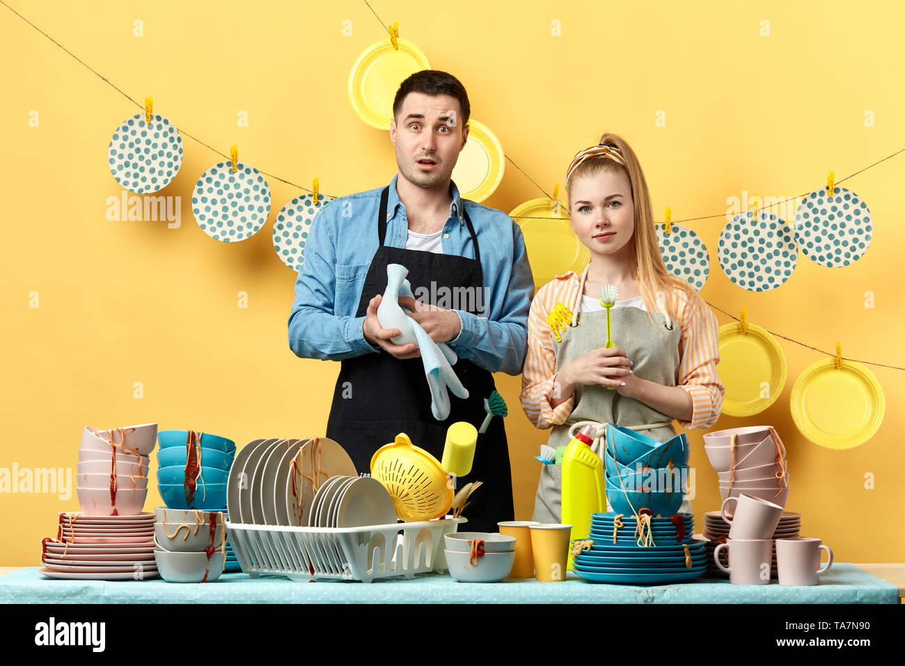 Schwerwiegende attraktive blonde Frau mit Pinsel und überrascht Mann mit einer Platte und einem Handtuch hinter dem Tisch und Blick in die Kamera. lifes Stockfoto