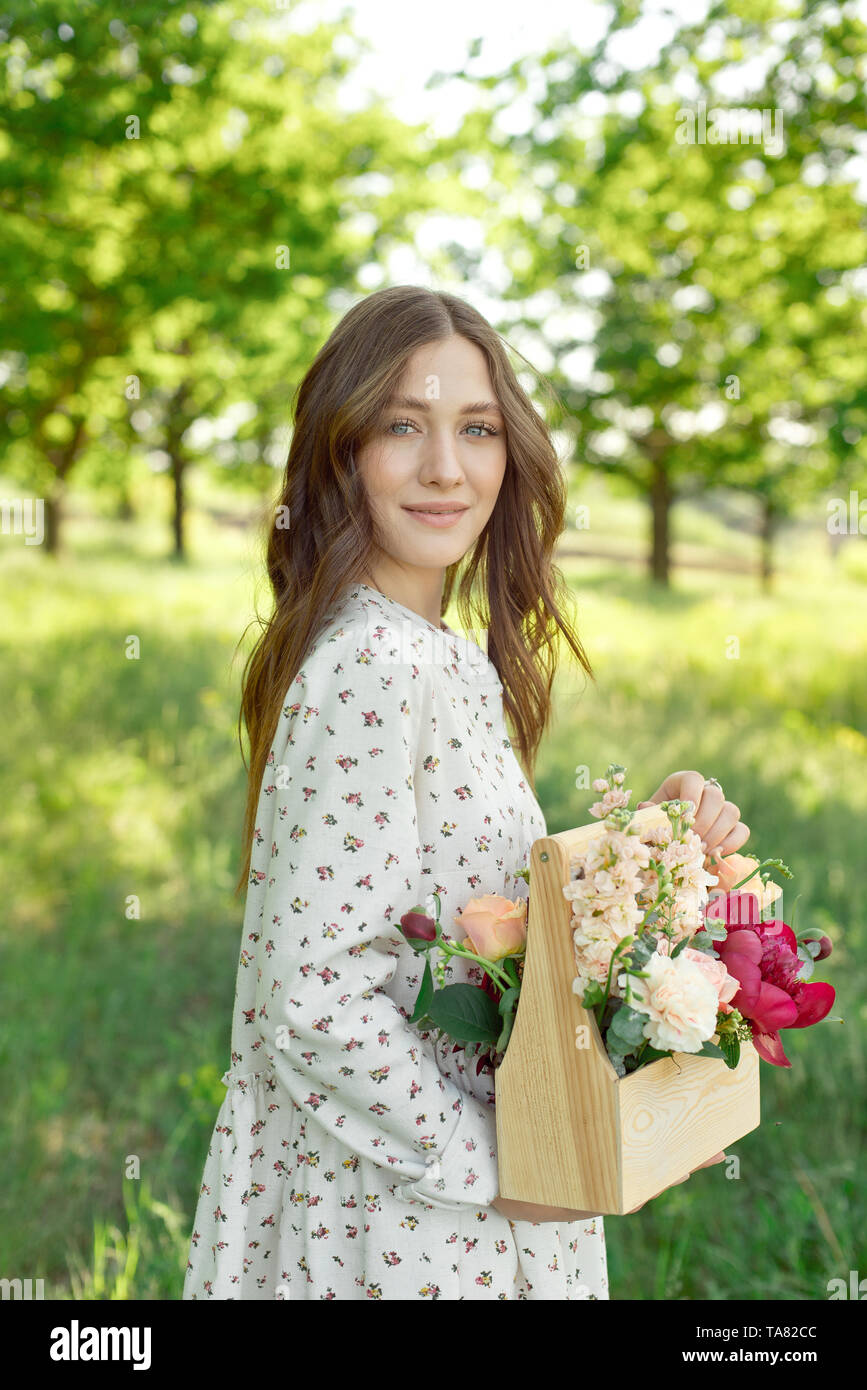 Die Hälfte Porträt eines charmanten positive Frau in langen weißen Sommerkleider mit einem glücklichen Lächeln auf dem Hintergrund einer grünen Wiese in den Händen der gekleidet Stockfoto