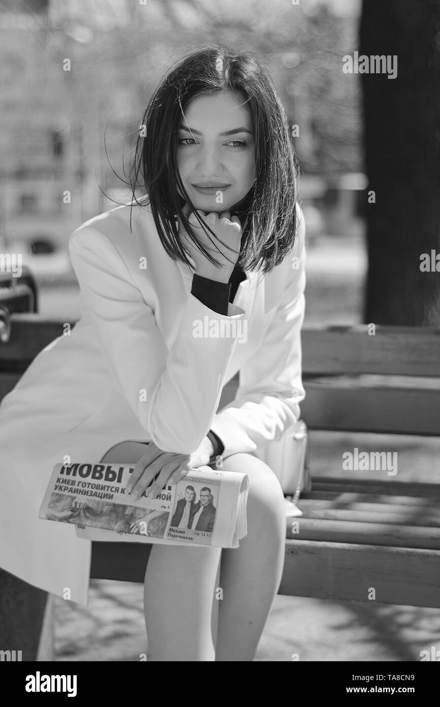 Elegante junge Frau in einem kurzen weißen Mantel auf einer Straße der Stadt mit einer Zeitung in ihren Händen posing gekleidet. Brünette Frau. Moderne urbane Frau Porträt. Stockfoto