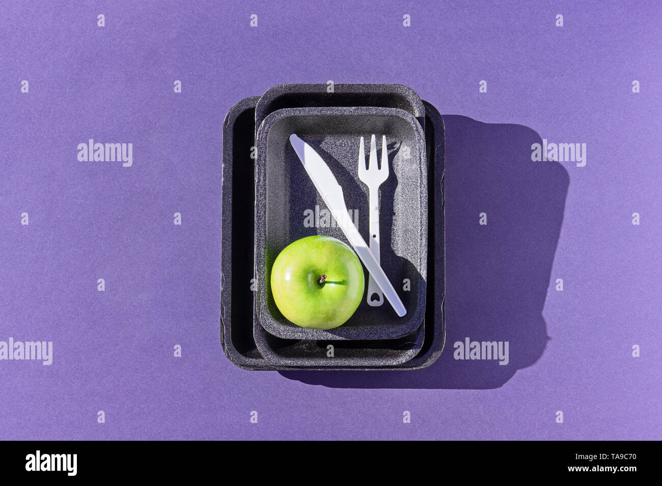 Blick von oben auf eine leere Kunststoff Fach mit grünem Apfel und Einweggeschirr auf lila, flach. Konzept der Verringerung der Verwendung von Kunststoff Stockfoto