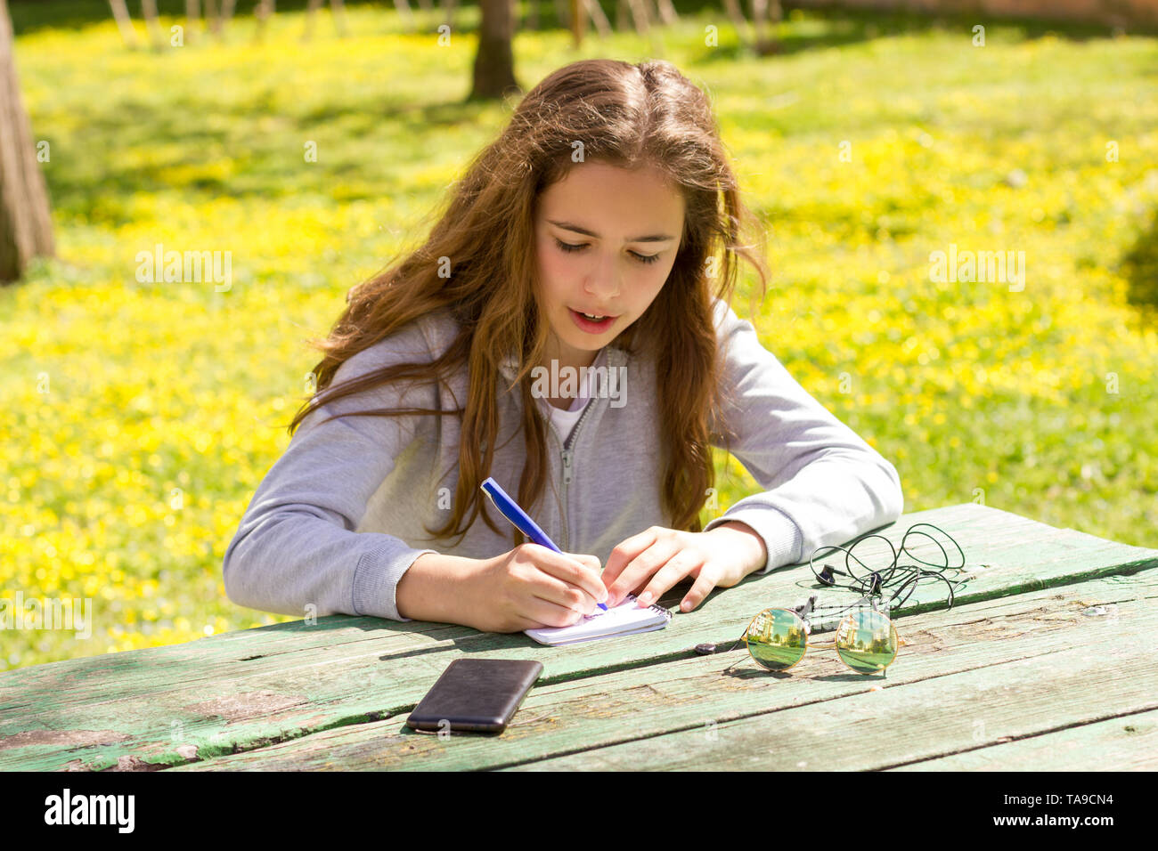 Cute Teen Girl schreibt Notizen auf einem Papierblock durch hölzerne Tabelle auf der grünen Wiese Stockfoto