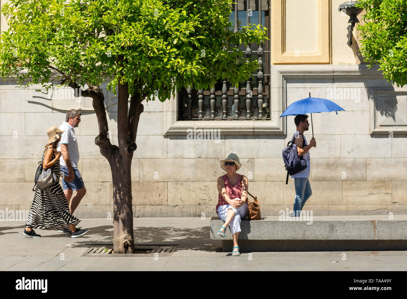 Frau sitzen auf einer Bank in Sevilla, Andalusien, Spanien. Ein Mann mit einem blauen Sonnenschirm zu Fuß durch. Stockfoto