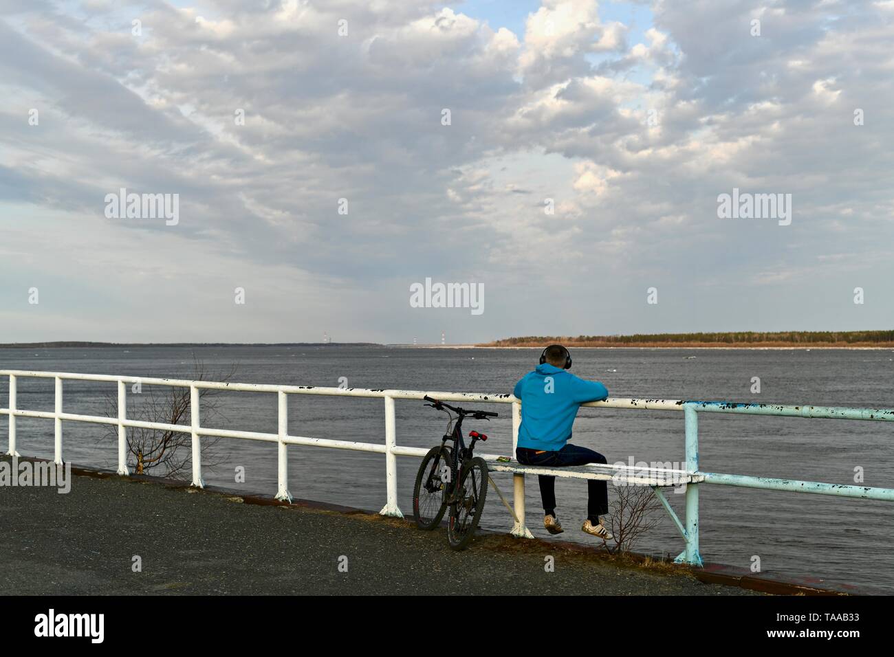 Surgut, Khanty-Mansiyskiy avtonomnyy Okrug, Rossiya - 10. Mai 2019: Damm des Ob-Fluss. Die ice Drift zu einem Ende kommt. Wolken schweben niedrig und der Himmel wird eine Reflexion auf den Fluss. Nach einem langen Winter, Bewohner des Nordens endlich ein Wechsel der Jahreszeit erlebt. Ein junger Mann mit Kopfhörern sitzt auf der Bank der Damm und in die Ferne schaut auf den Fluss, auf die Horizontlinie, wo der Fluss Himmel treffen. Sein Fahrrad in der Nähe geparkt. Stockfoto