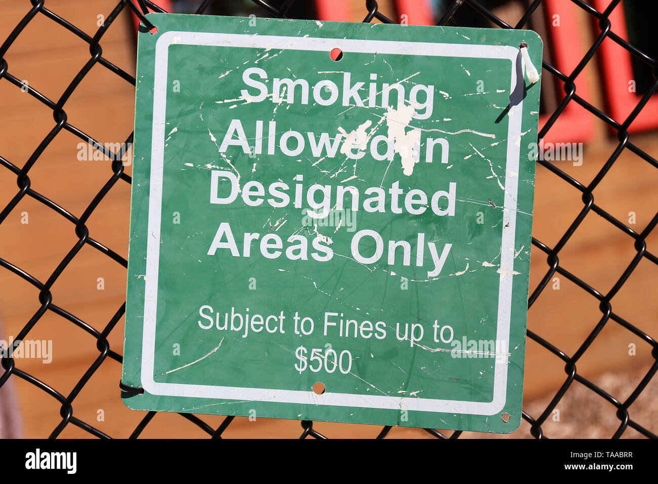 Ein grünes Schild ist auf einem Maschendrahtzaun auf einem Spielplatz gehangen, und sagt "Rauchen ist nur in den gekennzeichneten Bereichen erlaubt, $ 500 fein'. Stockfoto