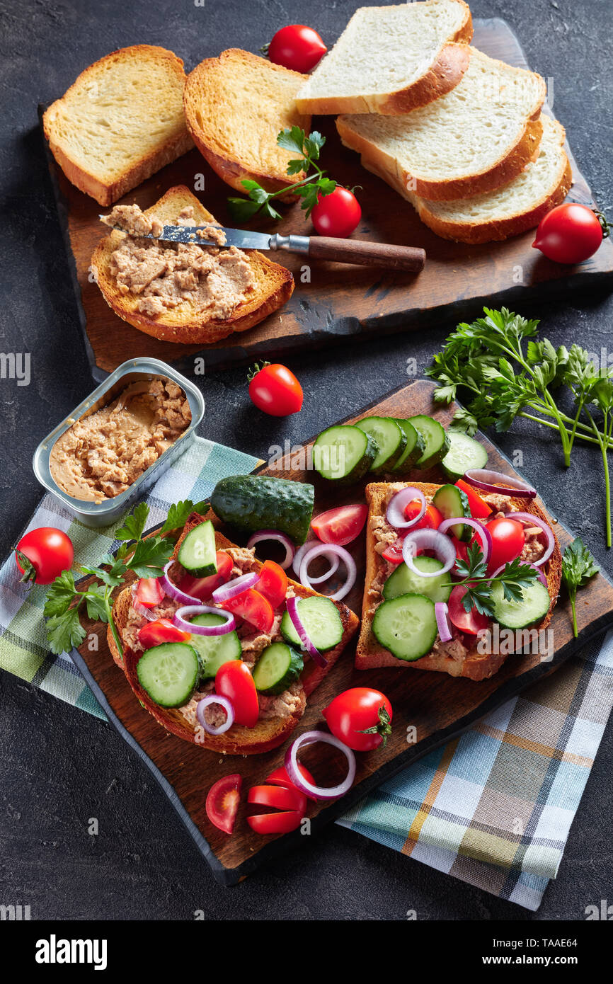 Toast für Sommer Frühstück mit Lebertran Pastete, gekrönt mit in Scheiben geschnittenen Gurken, Tomaten und roter Zwiebel Ringe auf einem Schneidebrett mit Zutaten aus einem Ki Stockfoto