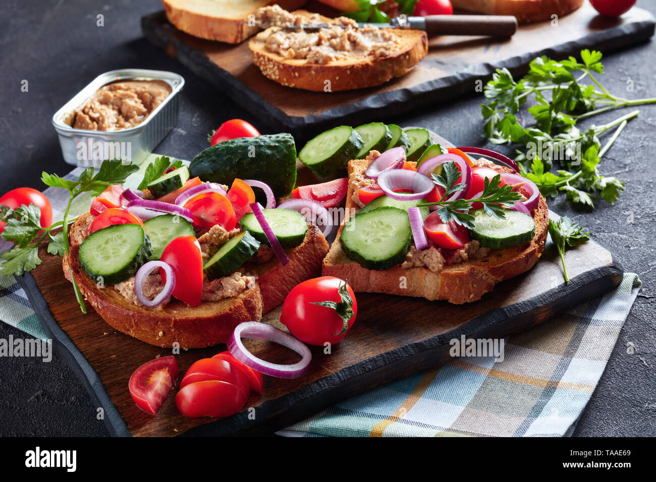 Toast mit Lebertran Creme, mit geschnittenen Gurken, Tomaten und roter Zwiebel Ringe auf einem Schneidebrett mit Zutaten auf einem Küchentisch überstieg, Horizon Stockfoto