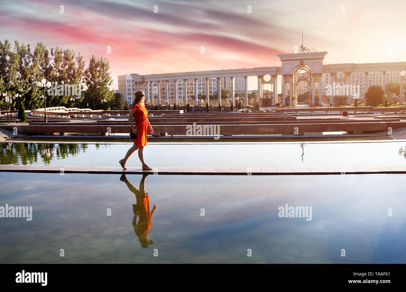 Frau in orange Kleid zu Fuß in der Nähe von Brunnen und Arch bei Sonnenaufgang in Almaty, Kasachstan Stockfoto