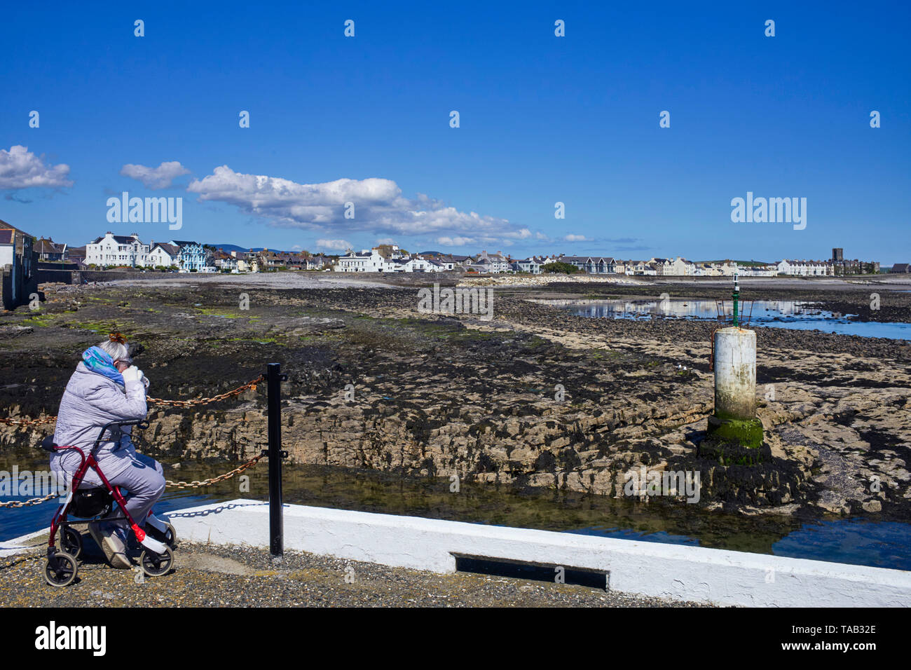 Ältere Frau tragen Steppjacke sitzen an der Seite des äußeren Hafenmauer in Castletown, Insel Man mit Blick auf die Bucht bei Ebbe Stockfoto