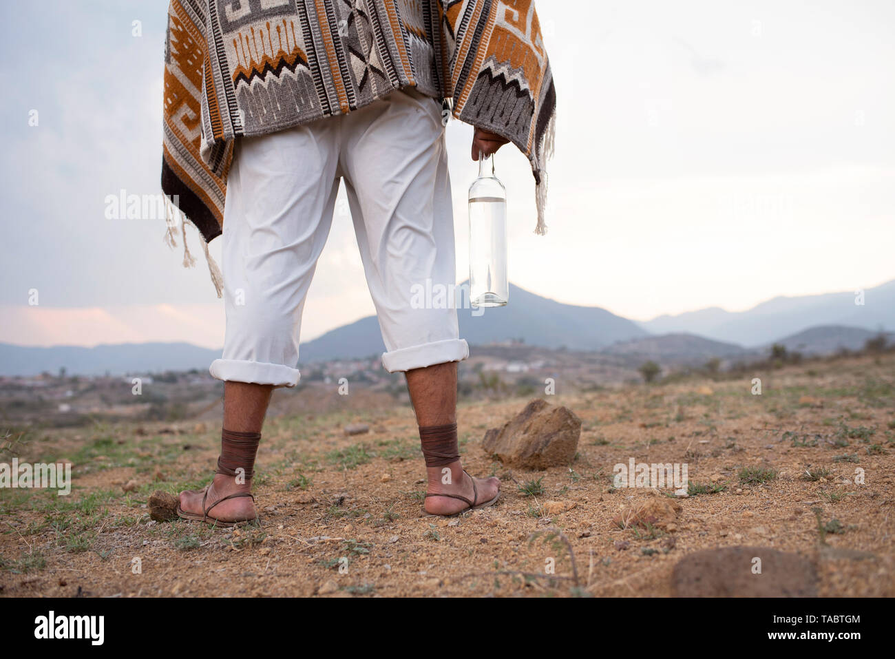 Mann von hinten stehend auf trockenem Boden tragen weiße Hosen, Zapotekisch Wolldecke, alte Sandalen und eine Flasche Mezcal. Oaxaca, Mexiko Stockfoto
