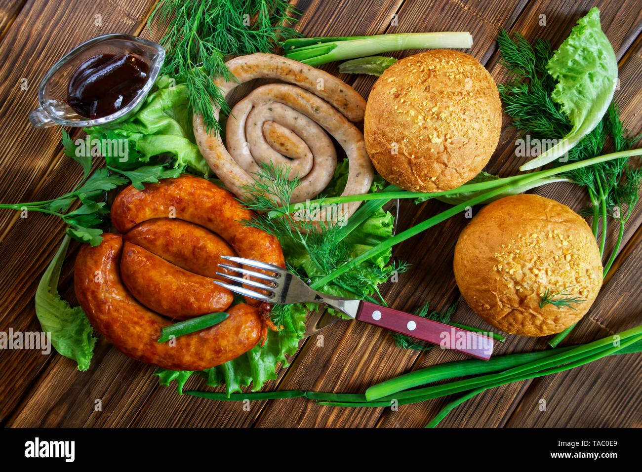 Mais Brötchen, gegrillte Würstchen, und eine transparente Sauce - pan liegen auf einem Holz- Oberfläche mit Gruenen: Salat, Dill, Zwiebel und Petersilie. Picknick - Grill. Vie Stockfoto
