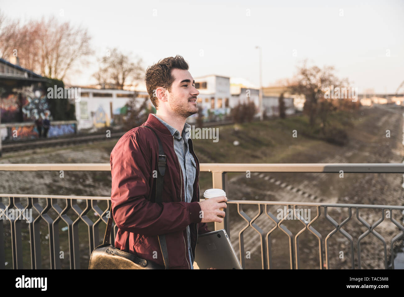 Junger Mann mit Tablet, Tasche und Kaffee zum Mitnehmen auf einer Brücke in der Stadt Stockfoto