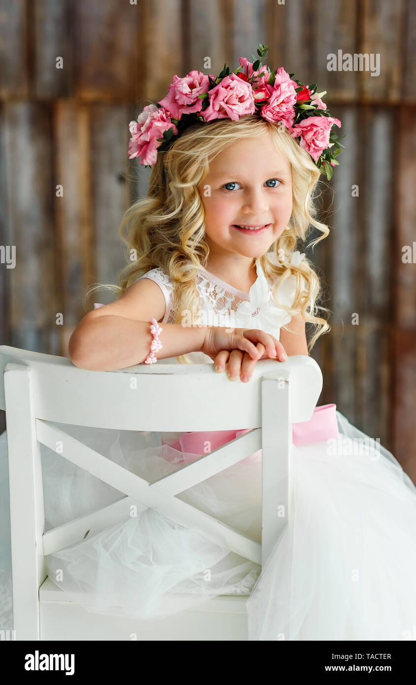 Schöne süße kleine Mädchen - Blondine in einem Kranz von Rosen in einem weißen Kleid in einem hellen Studio Stockfoto