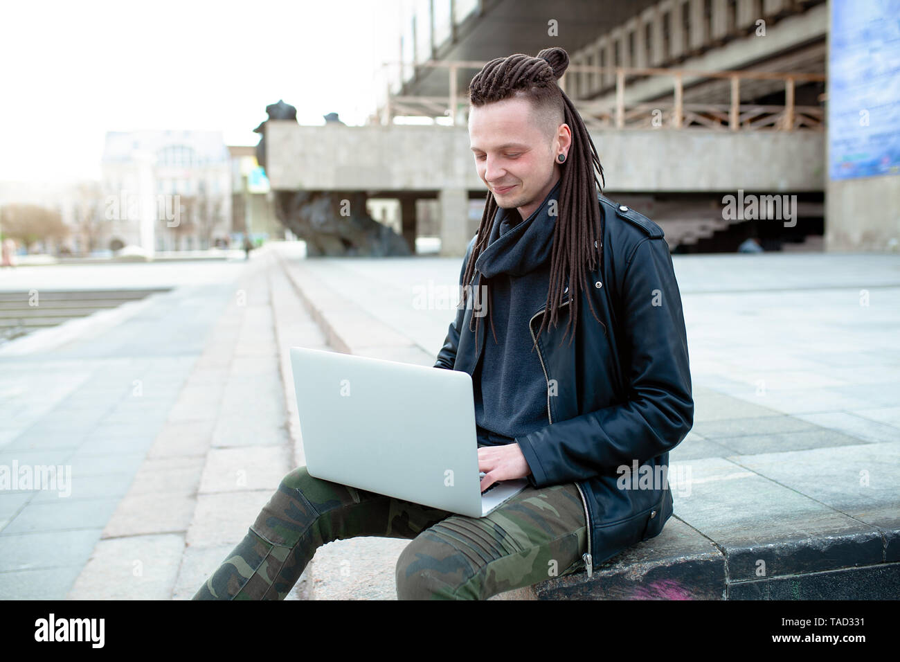Junge modische kaukasischen Mann mit Dreadlocks in Leder Jacke sitzen auf der Treppe mit Laptop gekleidet. Remote funktioniert IT-Spezialist Stockfoto