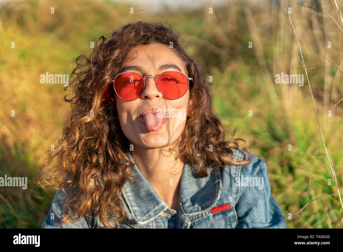 Porträt der jungen Frau mit dem lockigen braunen Haar zu tragen rote Sonnenbrille heraus haften Zunge Stockfoto
