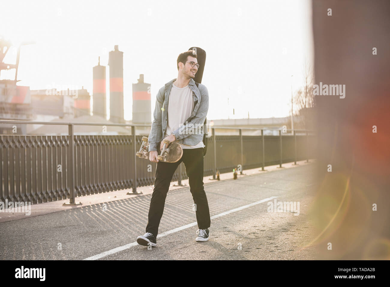 Lächelnden jungen Mann mit Gitarre und Skateboard zu Fuß auf einer Brücke in der Stadt Stockfoto