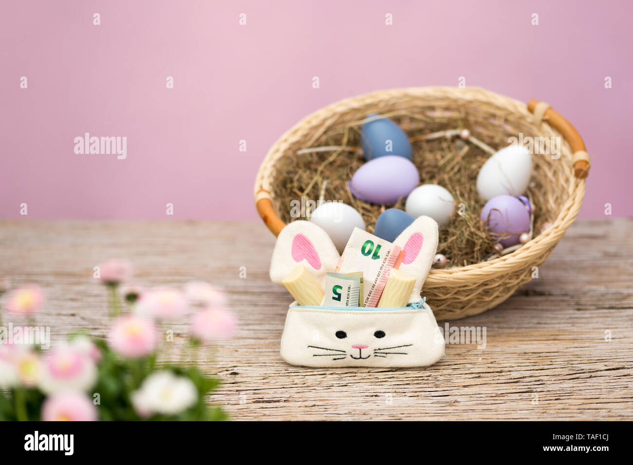 Eine kleine Tasche in der Form eines Hasen ist mit Geld und Süßigkeiten als Geschenk für Ostern gefüllt. Im Vordergrund sind Frühling Blumen, im Hintergrund Stockfoto