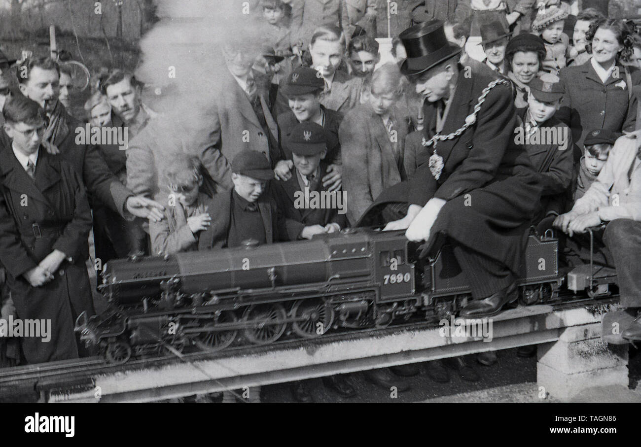 1950, historische, eine Masse von Menschen, einen älteren männlichen Würdenträger suchen, eine lolca Bürgermeister trägt einen Hut, Anzug und eine Kette, eine Fahrt in einem Dampfzug, auf eine Modelleisenbahn, England, UK. Diese enge gauage Miniatur-eisenbahn liegt über dem Boden und der Fahrer und Passagiere erhöhte sich rittlings auf die Spur. In dieser ära Modell oder Miniatur Eisenbahnen mit Dampflokomotiven waren eine beliebte Attraktion in Parks und Badeorten. Stockfoto