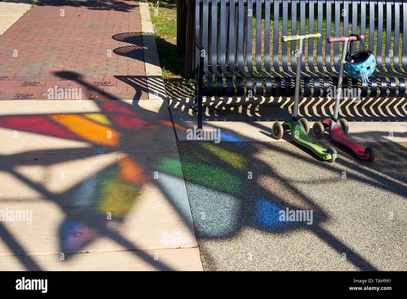 Zwei Kinder Scooter mit einem Helm rest Neben einer Parkbank mit einem interessanten Prisma Color Schattenwurf auf dem Beton Gehweg. Stockfoto