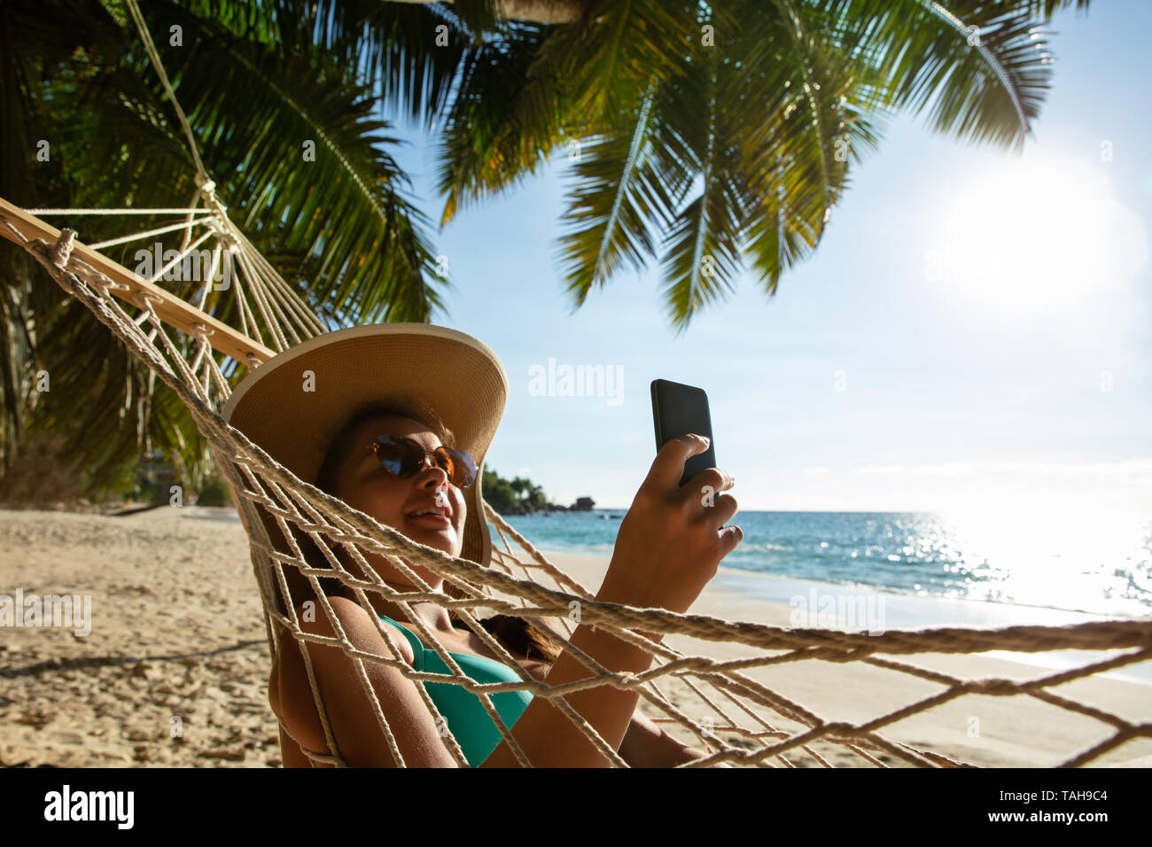 Lächelnden jungen Frau mit Hut entspannen auf der Hängematte mit Handy am Strand Stockfoto