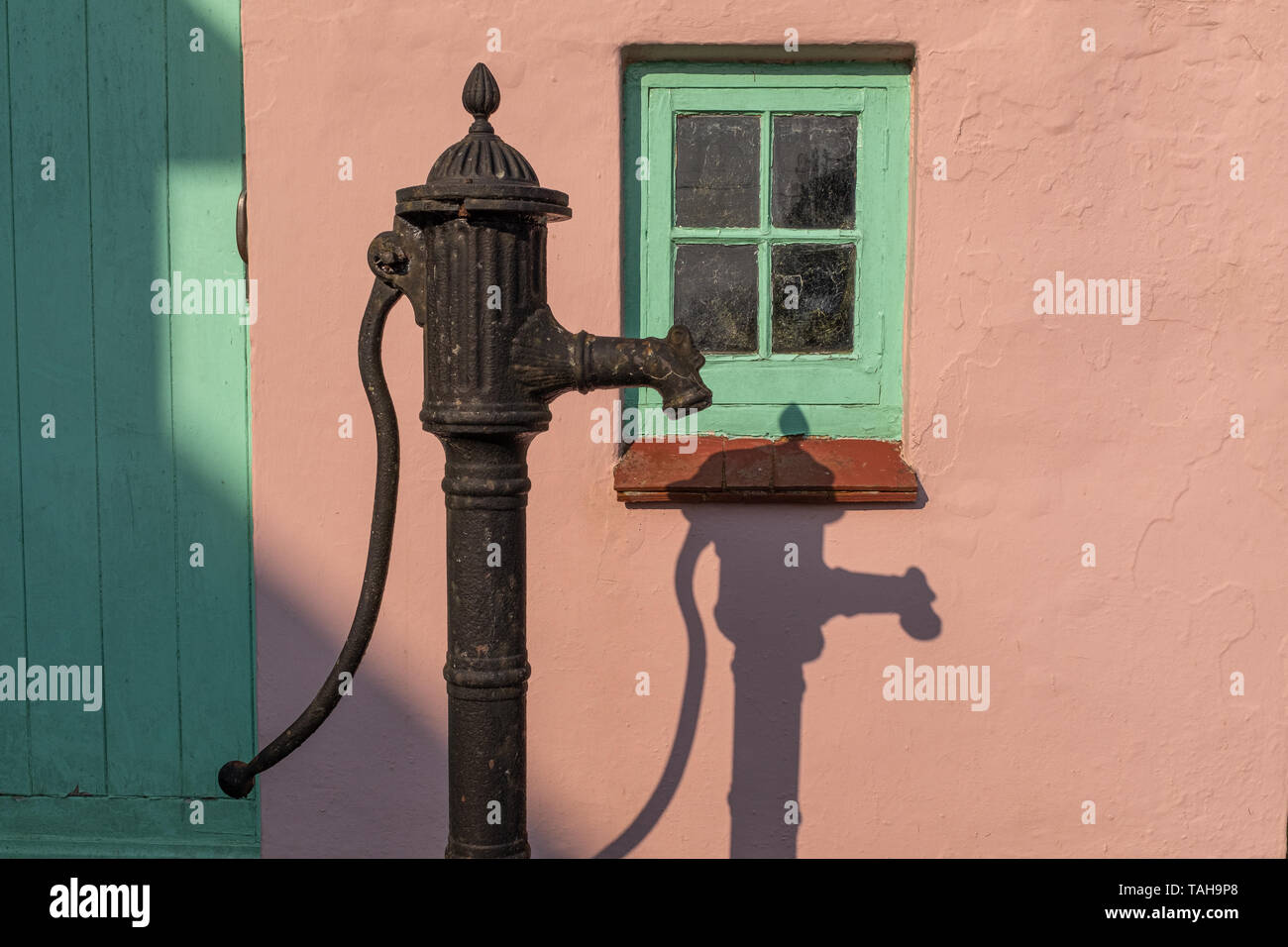 Eine altmodische Wasserpumpe gegen eine rosa Wand mit einem grünen Fenster, mit einem starken Schatten der Pumpe, niemand im Bild Stockfoto