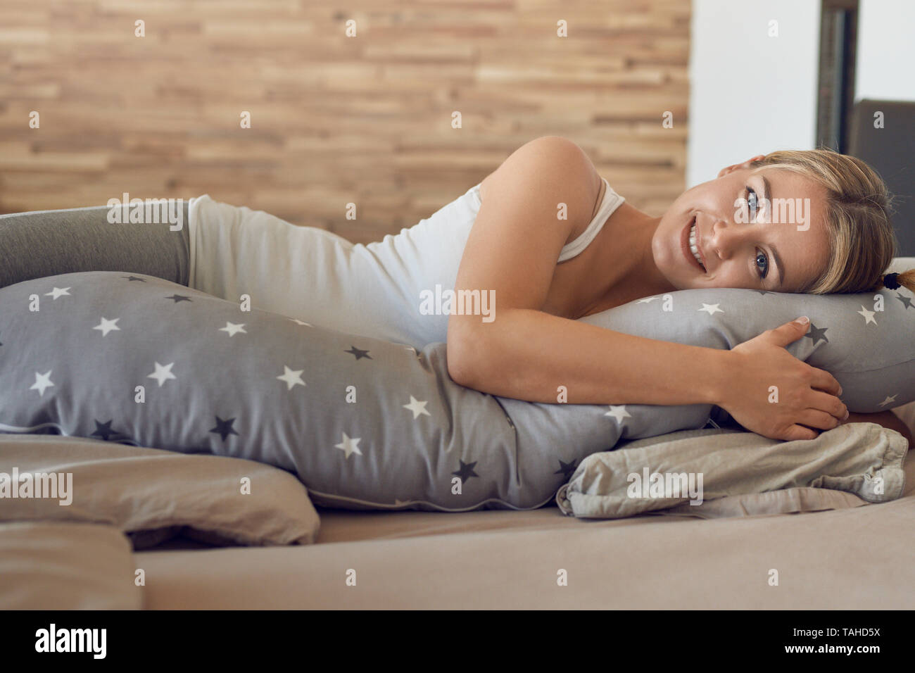 Glückliche gesunde junge schwangere Frau ruht auf einem Bett mit einem speziellen Kissen als Unterstützung in die Kamera schaut mit einem reizenden Lächeln Stockfoto